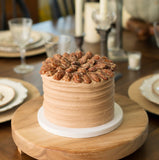 A round tiered cake with light tan frosting and a topping of chopped nuts, displayed on a wooden cake stand on a dining table surrounded by plates, candles, and pumpkins.