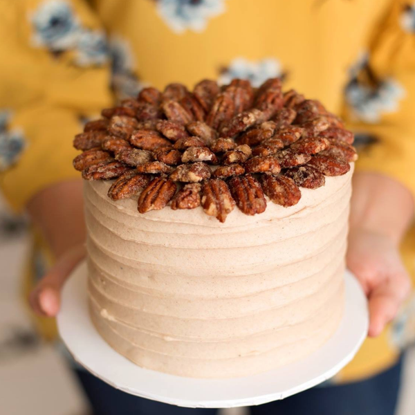 A round frosted sweet potato cake topped with a dense arrangement of whole pecans, held on a white plate by a person wearing a yellow floral top.